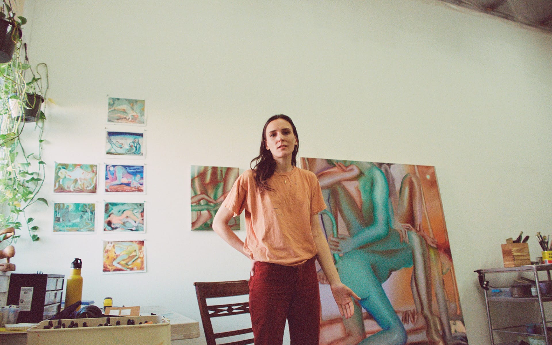 Artist standing in a studio with paintings on the wall, surrounded by art supplies and plants.