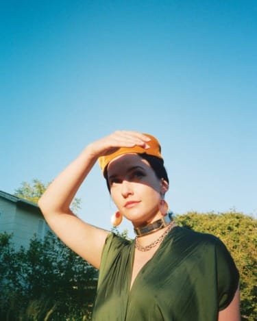 Person outdoors wearing a green outfit and yellow cap, shielding eyes from the sun with trees and blue sky in the background
