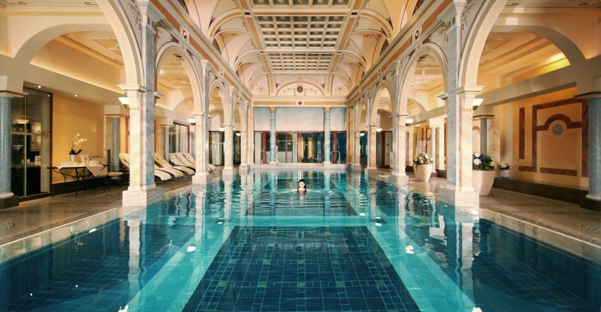 Luxurious indoor swimming pool with ornate arches, high ceilings, and a person swimming, surrounded by elegant lounge chairs.