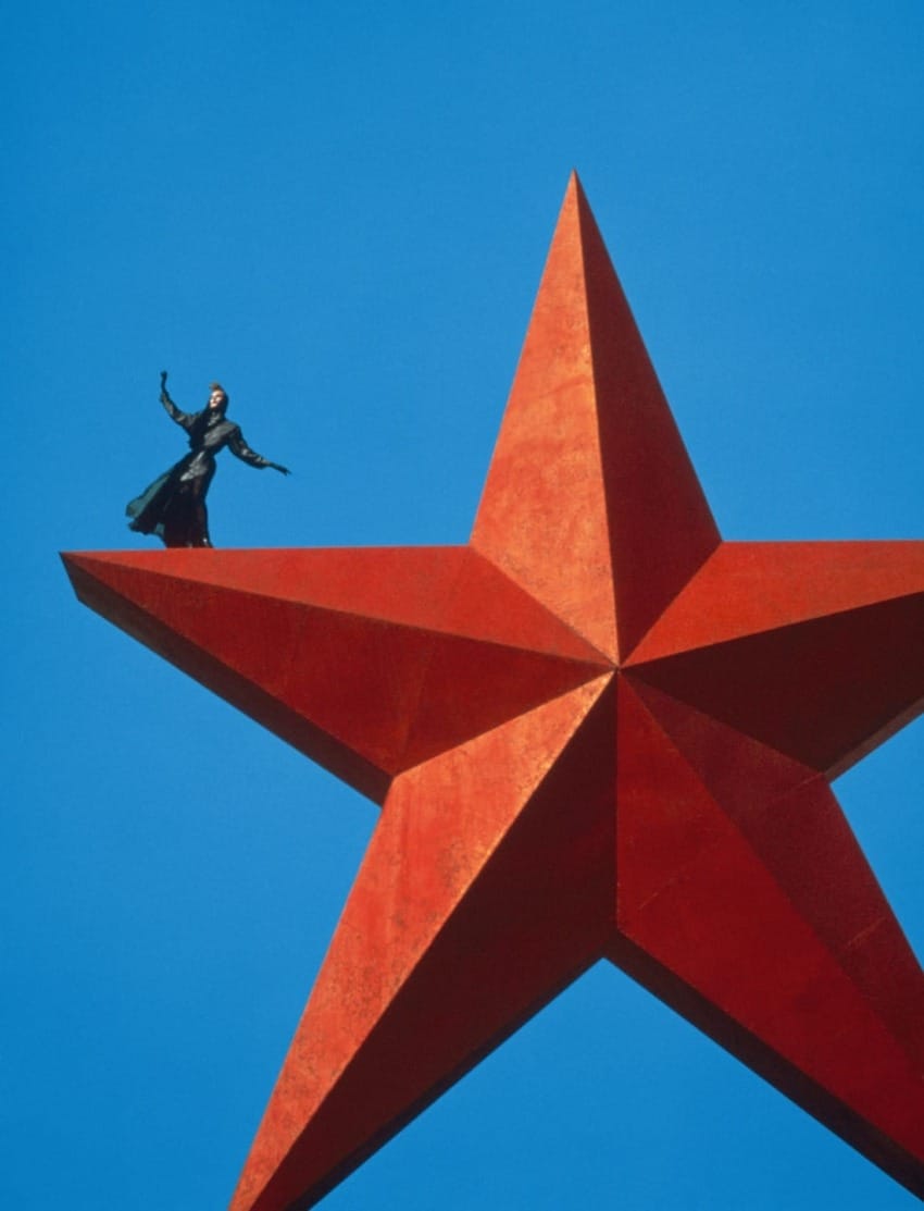 Person in dark attire standing on a large red star against clear blue sky backdrop.