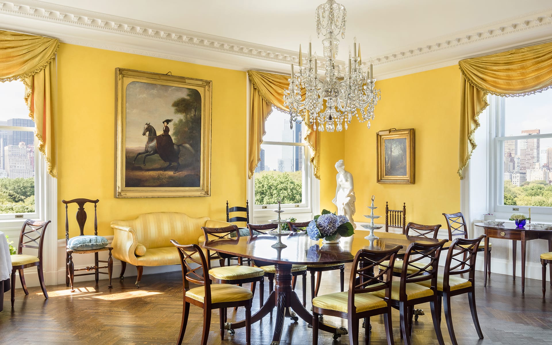 Elegant dining room with yellow walls, chandelier, large painting, and a view of trees through tall windows.