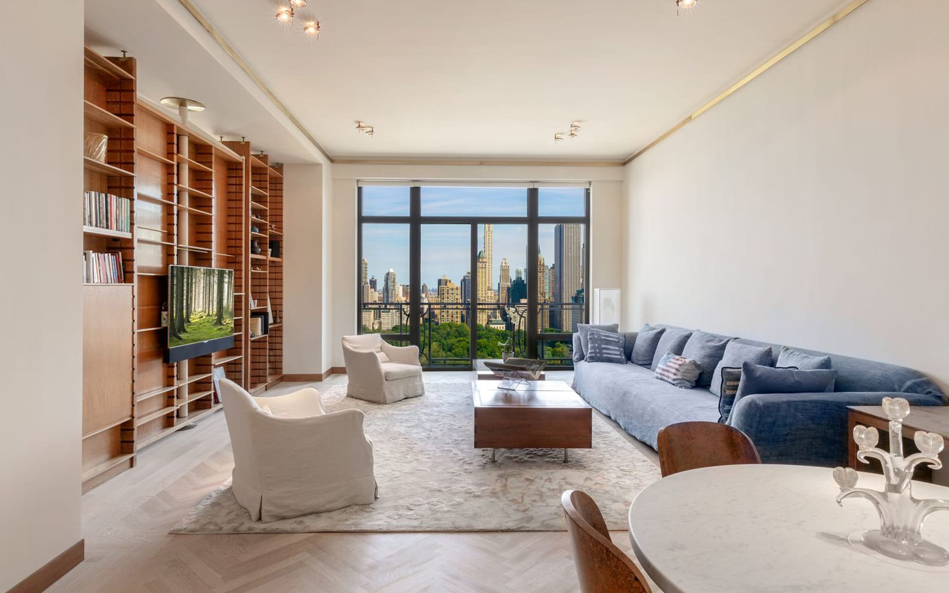 Modern living room with large windows, city skyline view, blue sofa, white armchairs, and wooden shelves.