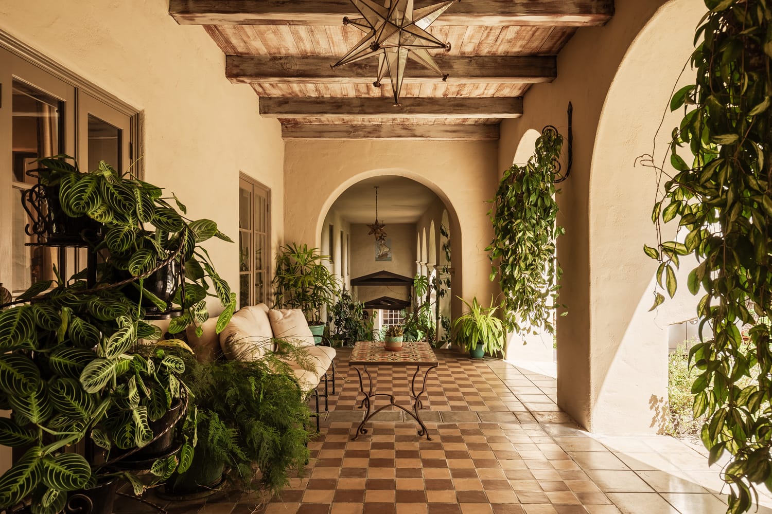 A rustic patio with potted plants, arched openings, a wooden ceiling, and a cushioned seating area on a checkered floor.