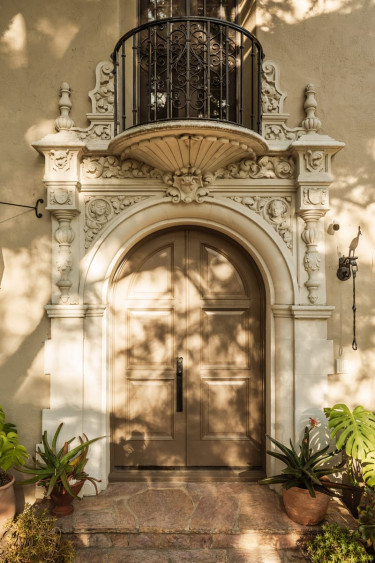 Ornate arched doorway with decorative stonework and small wrought iron balcony above surrounded by lush plants.