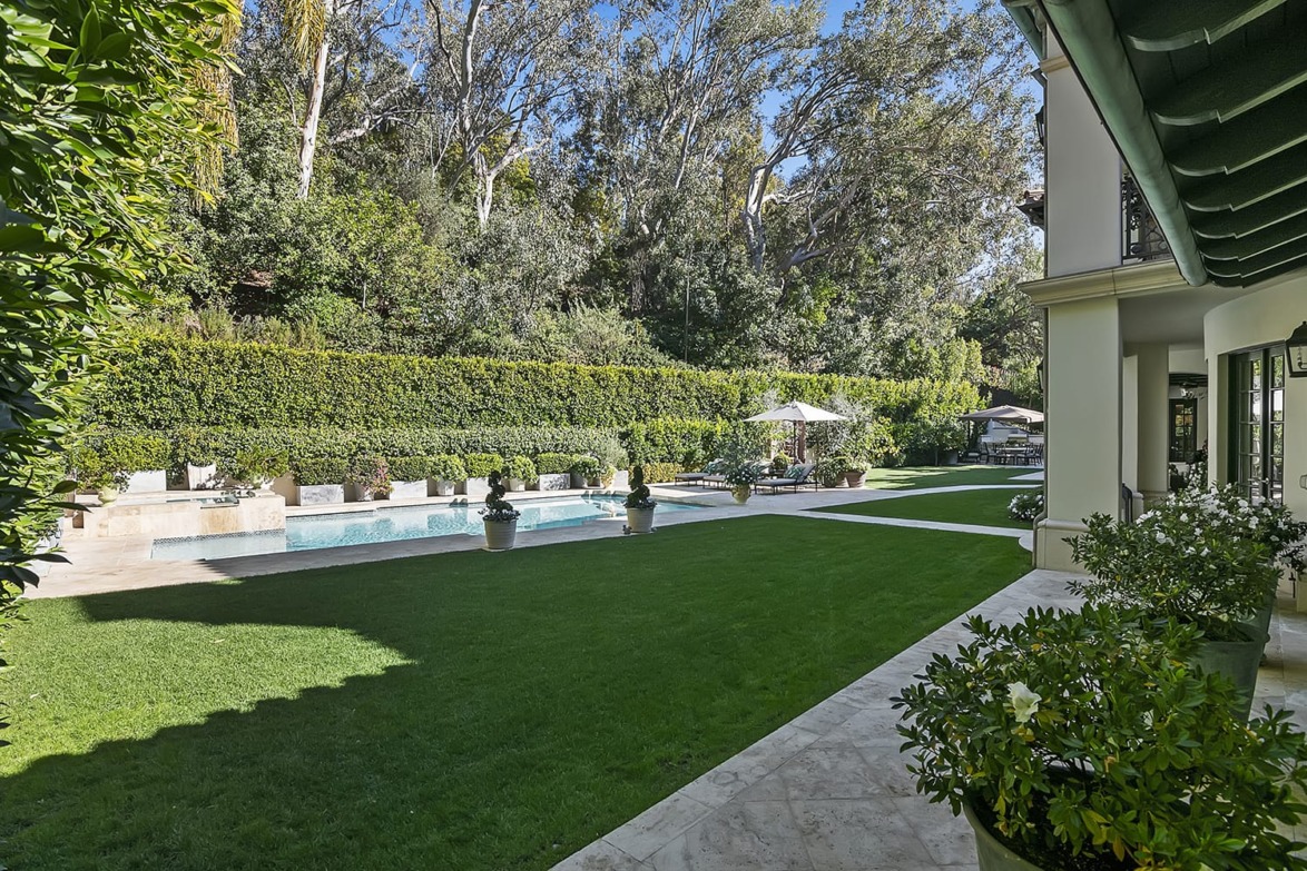 Luxurious backyard with a swimming pool, lush green lawn, trees, and a patio area on a sunny day.