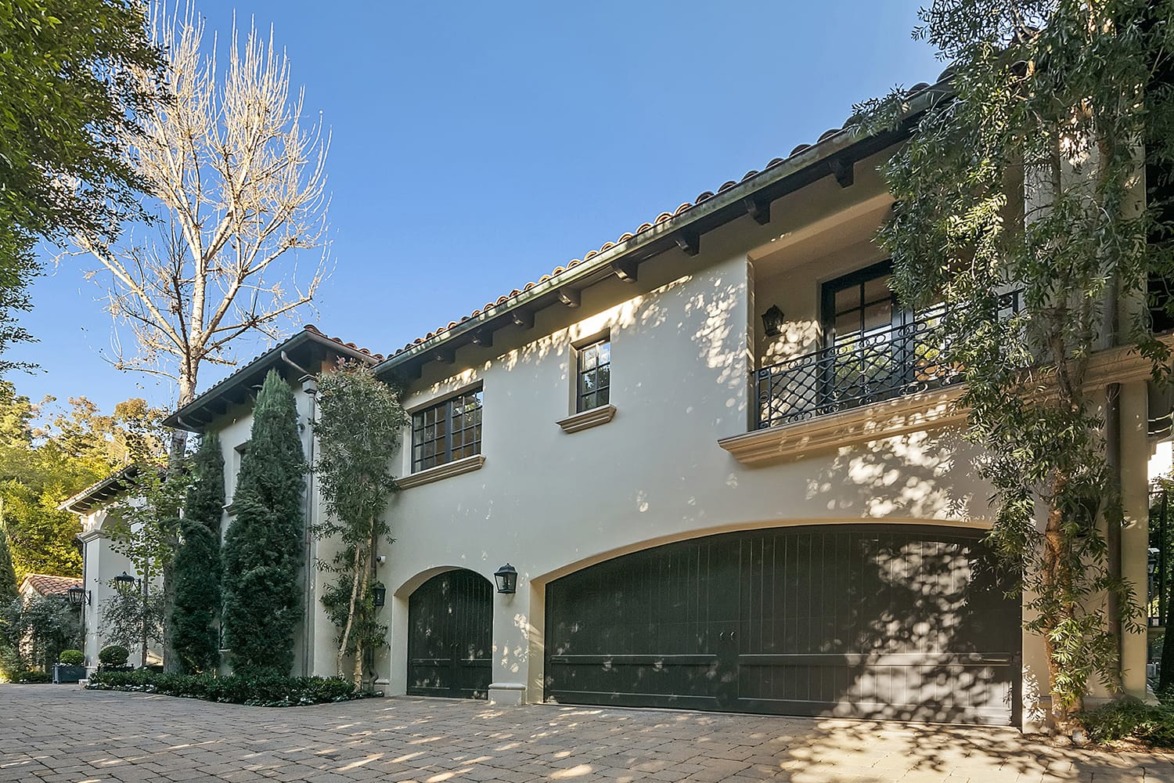 Mediterranean-style house with two-car garage, light stucco walls, and lush greenery surrounding the driveway.