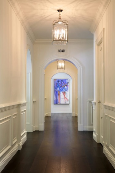 Elegant hallway with dark wood floor, white walls, decorative molding, and a modern chandelier, leading to colorful artwork.