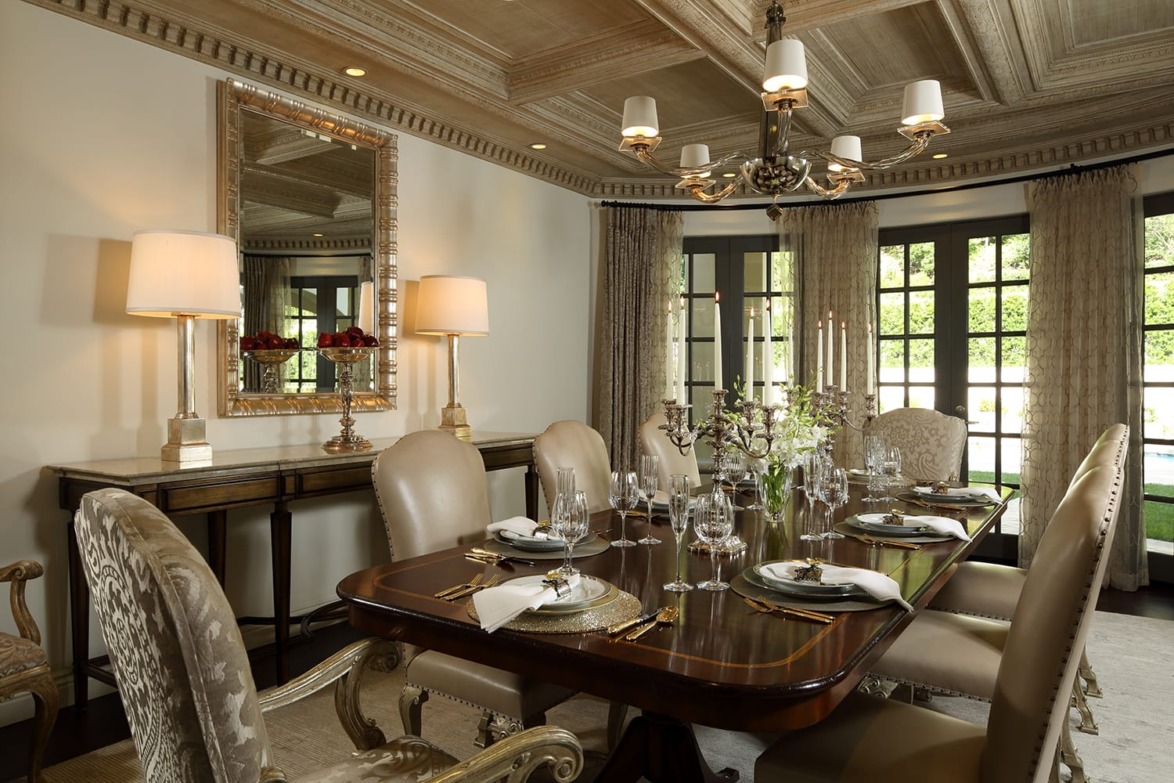 Elegant dining room with a wooden table set for dinner, luxurious chairs, chandelier, and large windows with curtains.