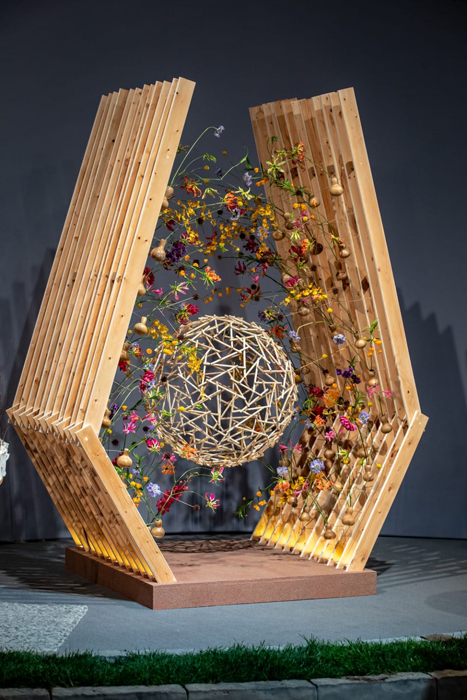 Wooden art structure with intricate sphere, adorned with colorful flowers and plants, displayed in an indoor setting.