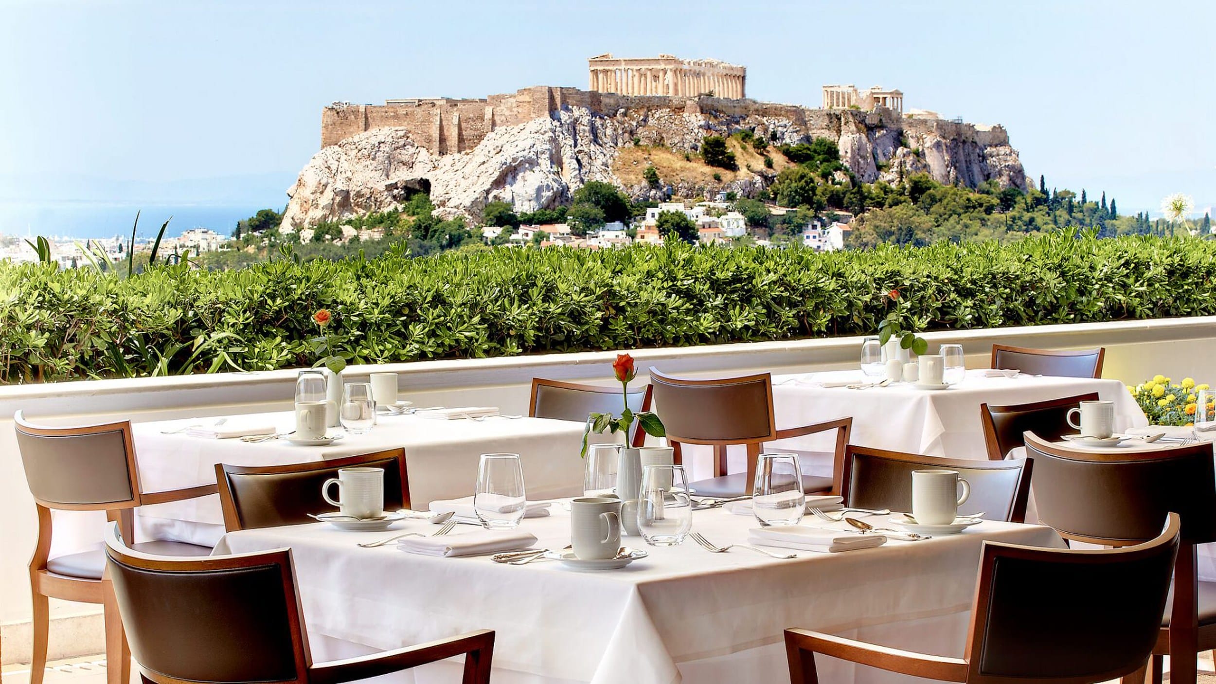 View of the Acropolis from a restaurant terrace with neatly set tables and chairs in the foreground.