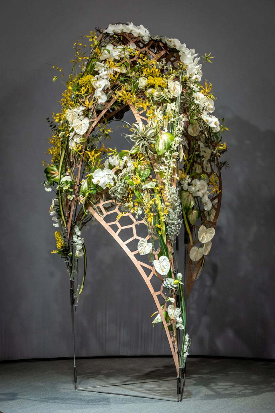 Floral arch sculpture with white and yellow flowers on a modern geometric wooden frame against a gray background.