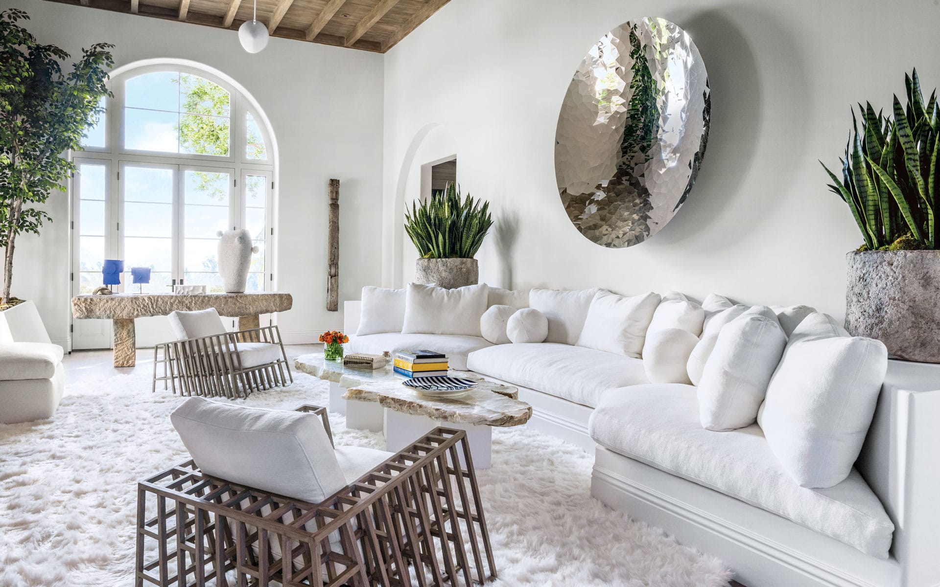 Modern living room with white furniture, abstract wall art, large windows, and indoor plants on a fluffy white rug.