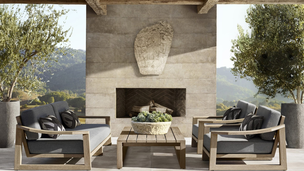 Outdoor patio with modern wooden furniture, gray cushions, potted plants, and a stone fireplace under a wooden pergola.
