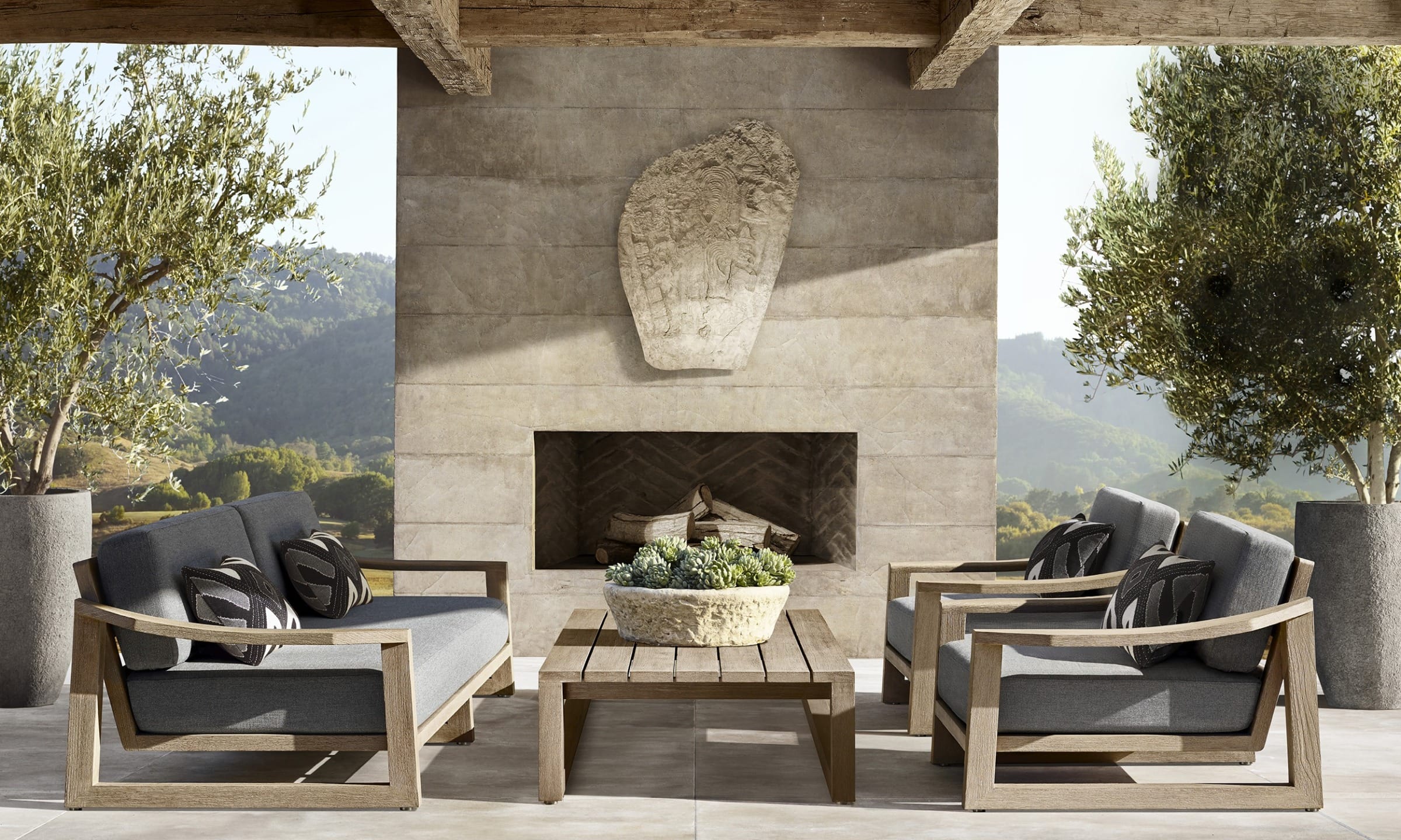 Outdoor patio with modern wooden furniture, gray cushions, potted plants, and a stone fireplace under a wooden pergola.