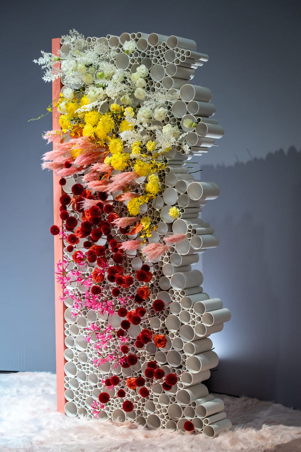 Colorful floral arrangement in white cylindrical tubes, featuring red, yellow, pink, and white flowers on a textured base.