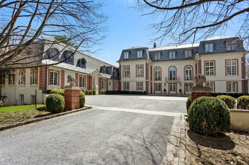 Large elegant mansion with a courtyard, surrounded by trees and bushes under a clear blue sky.