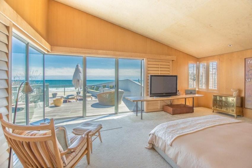 Bright beachfront bedroom with large windows, ocean view, wooden furniture, and cozy decor.