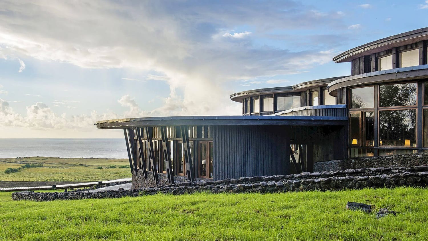 Modern building with large windows overlooking a grassy landscape and ocean under a partly cloudy sky.
