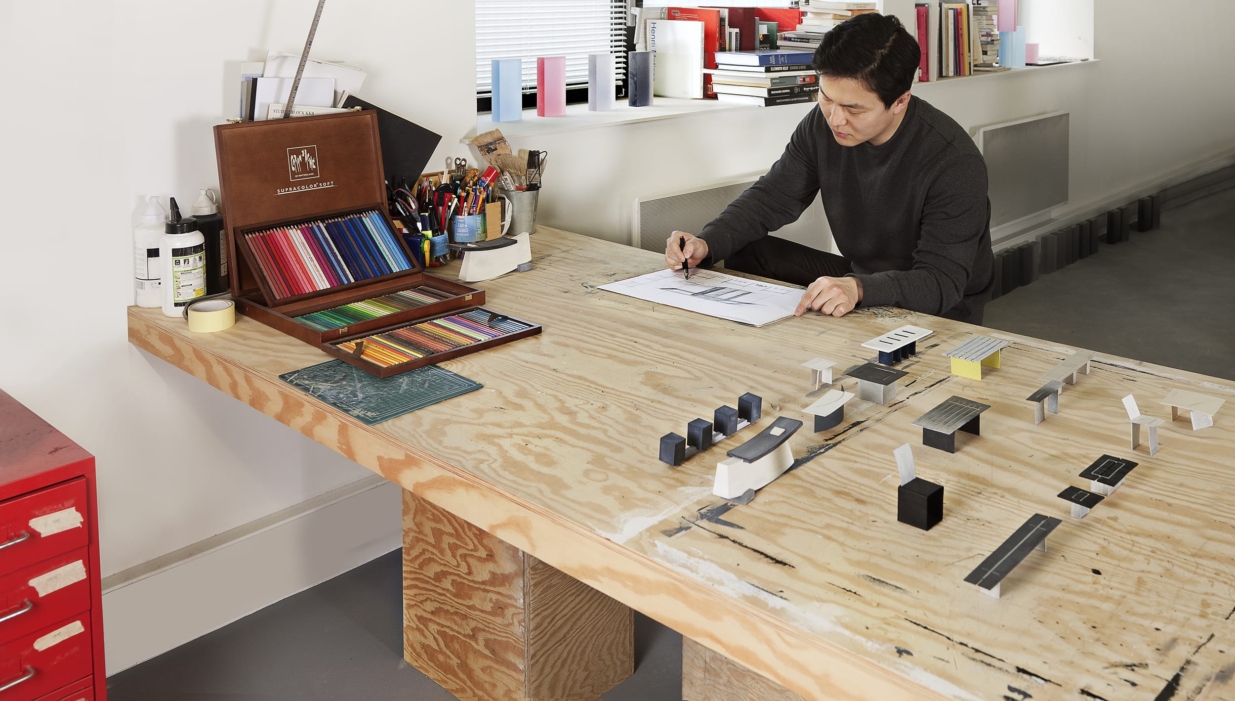 Man sitting at a large wooden desk working on architectural designs with colored pencils and scale models scattered around.