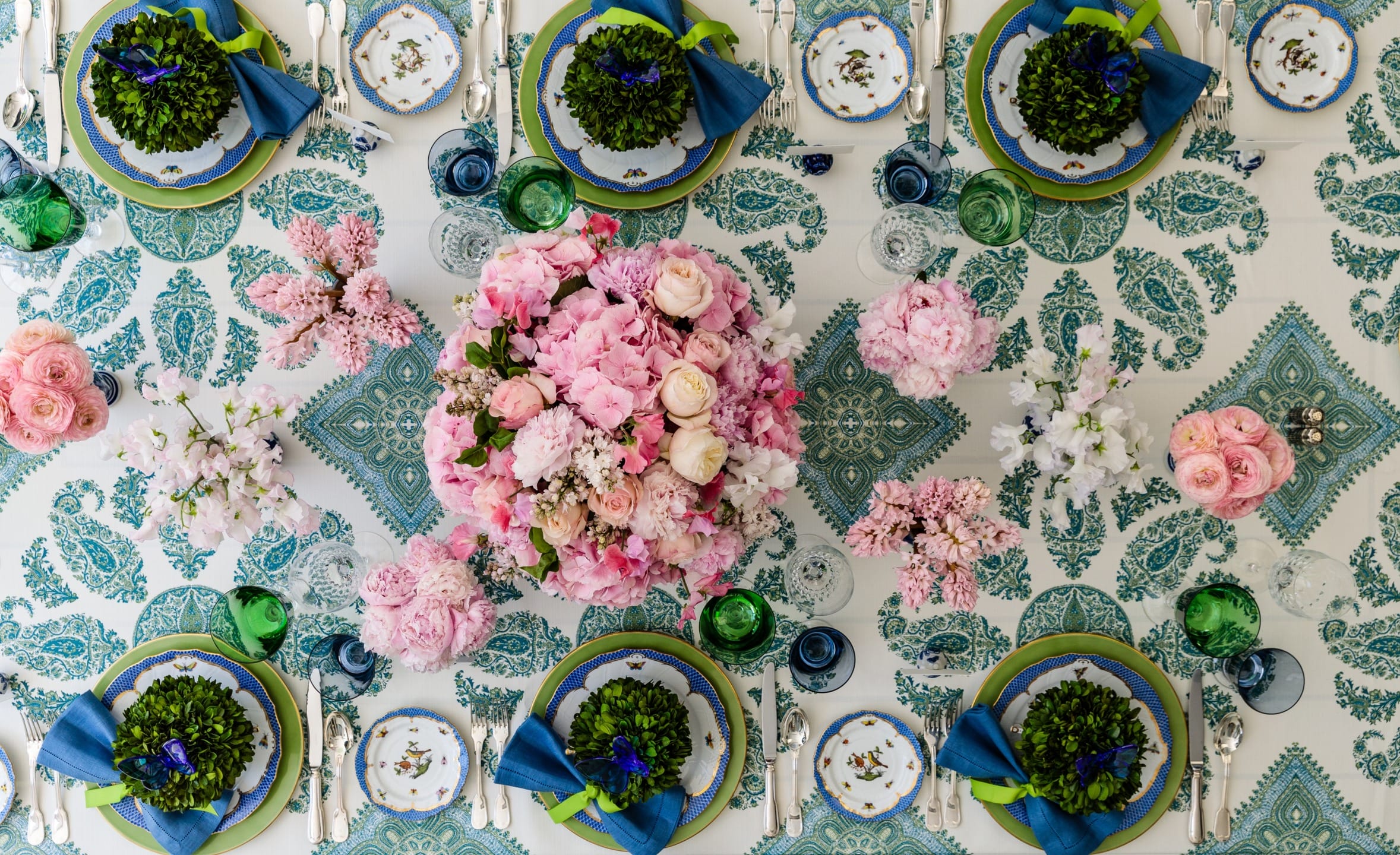 Overhead view of a vibrant, floral table setting with pink flowers, blue accents, and leafy green decorations.