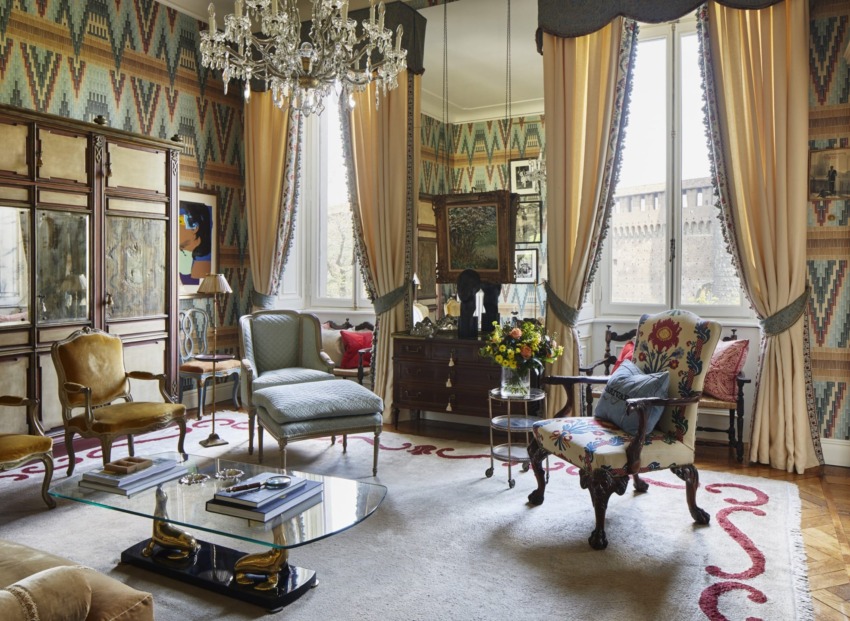 Elegant living room with vintage furniture, crystal chandelier, patterned wallpaper, and large windows with drapes.