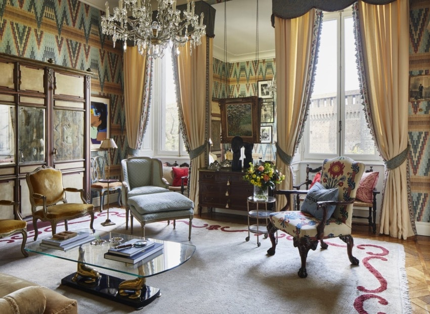 Elegant living room with vintage furniture, crystal chandelier, patterned wallpaper, and large windows with drapes.