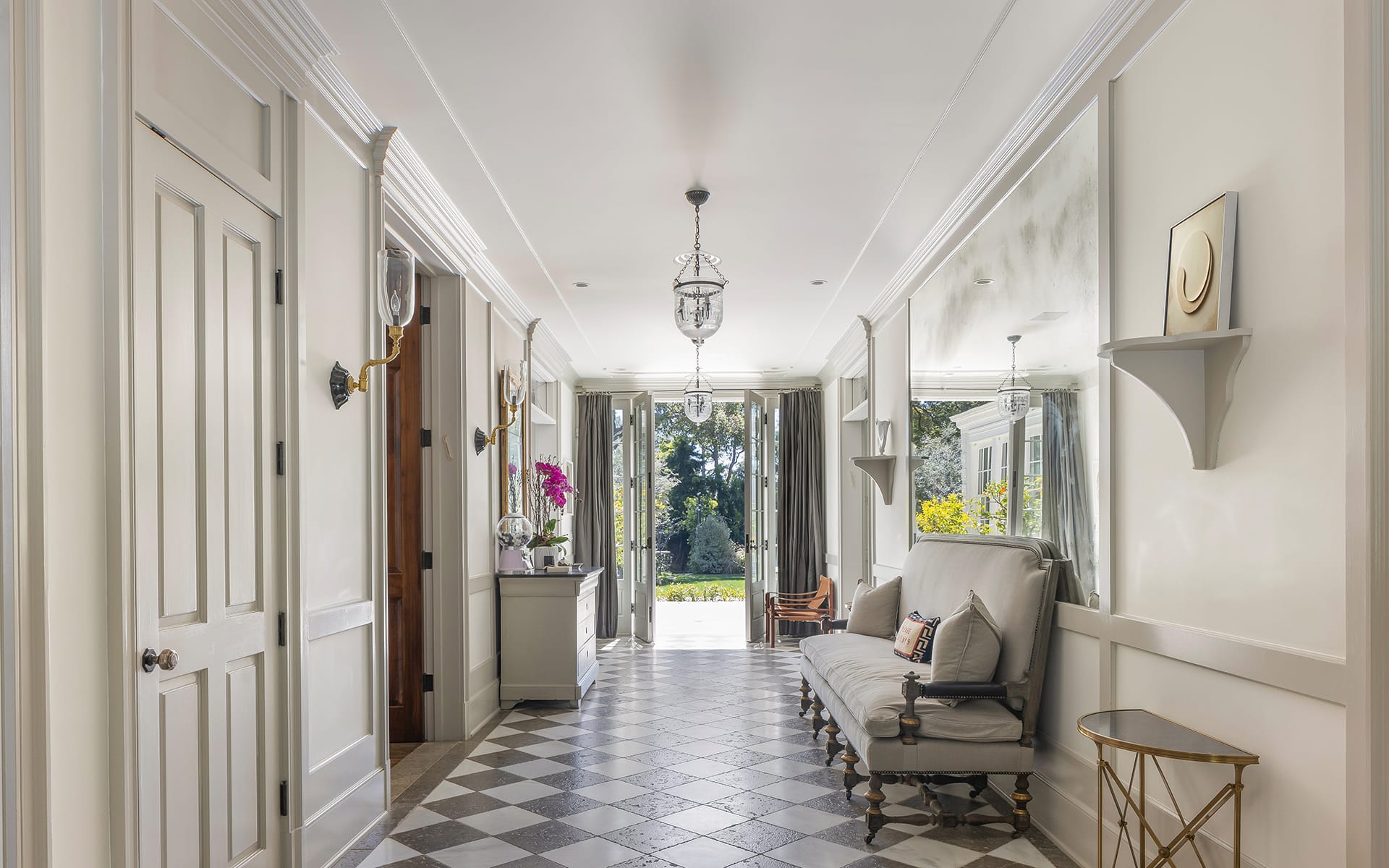 Elegant hallway with checkered floor, wall sconces, and cozy seating, leading to open doors revealing a garden view.