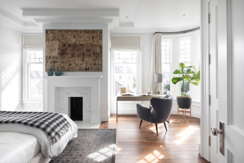 Bright bedroom with a modern chair, desk, and potted plant beside a fireplace, featuring large windows and hardwood floor.