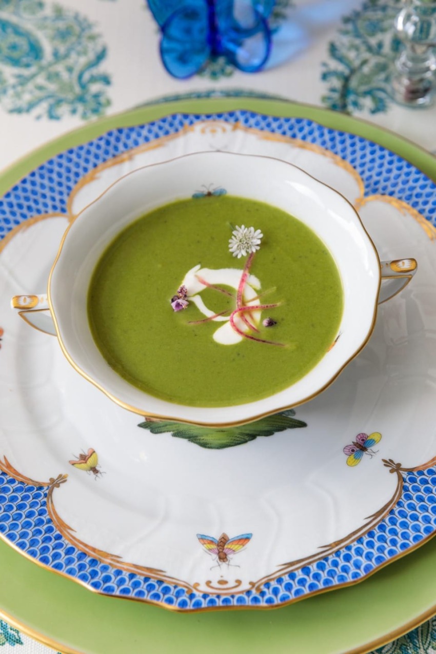 Elegant bowl of green soup garnished with edible flowers on a patterned plate with blue accents.