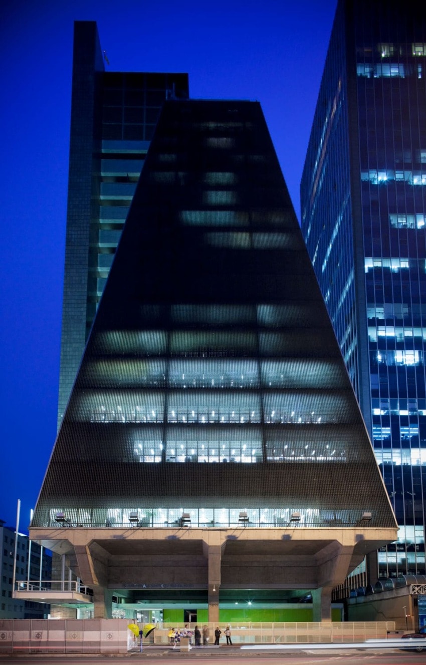 Pyramidal modern skyscraper at night with illuminated windows, towering over a city street with people walking below.