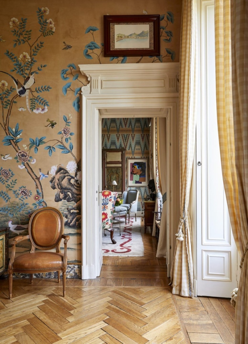 A room with ornate wallpaper, wooden flooring, and a view into another room with eclectic decor and patterned textiles.
