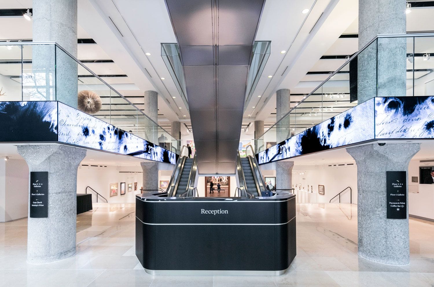 Modern lobby with escalators, large columns, digital displays above, and a central reception desk in a spacious interior.