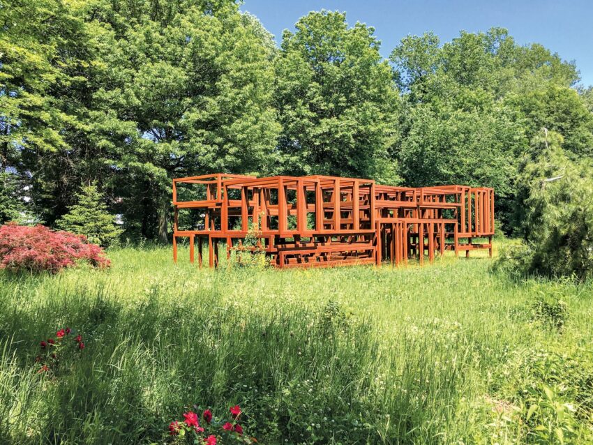 Red metal sculpture in a green meadow surrounded by trees, with red flowers in the foreground on a sunny day.