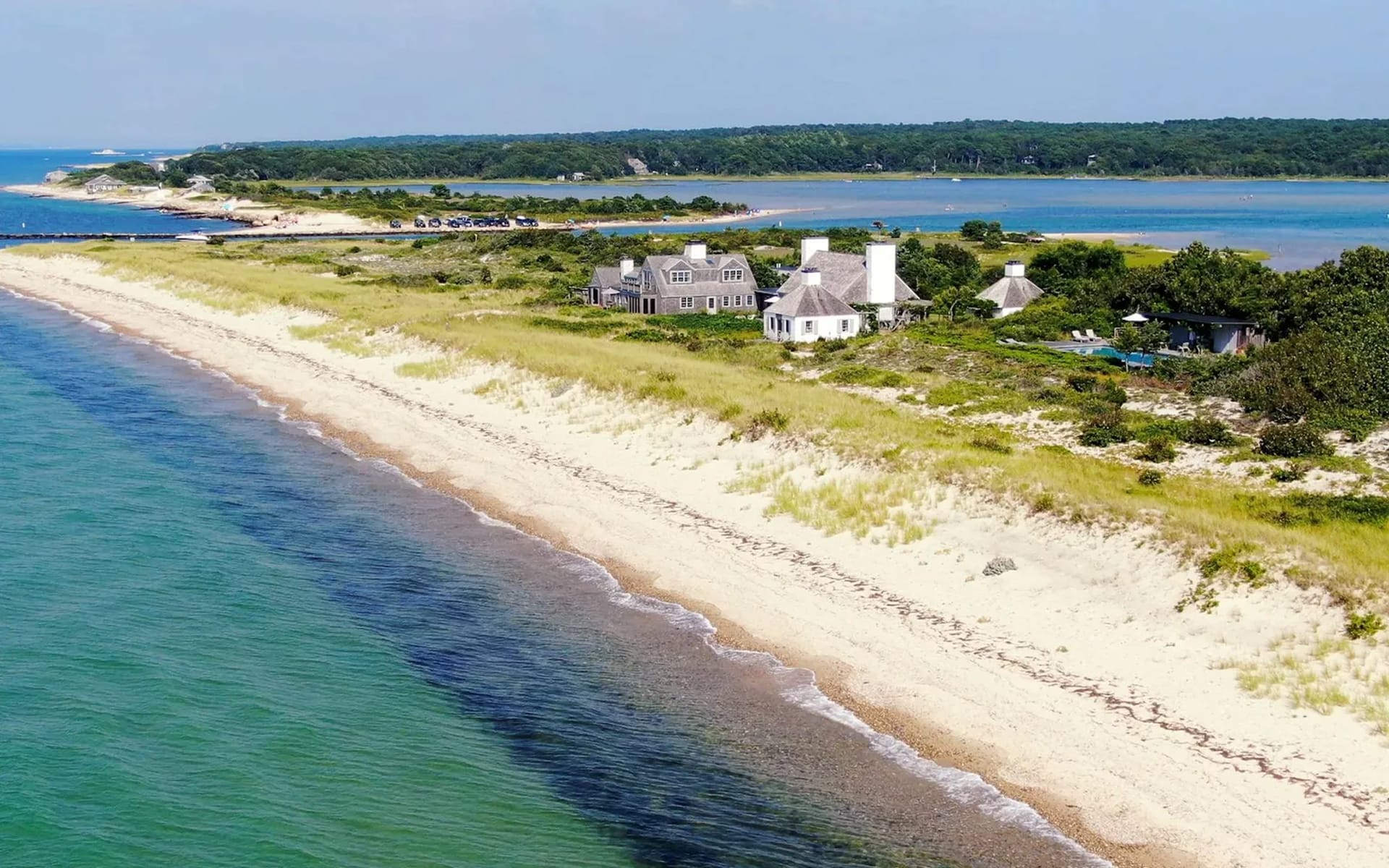 Aerial view of a coastal landscape with sandy beach, lush greenery, and houses by the ocean under a clear blue sky.
