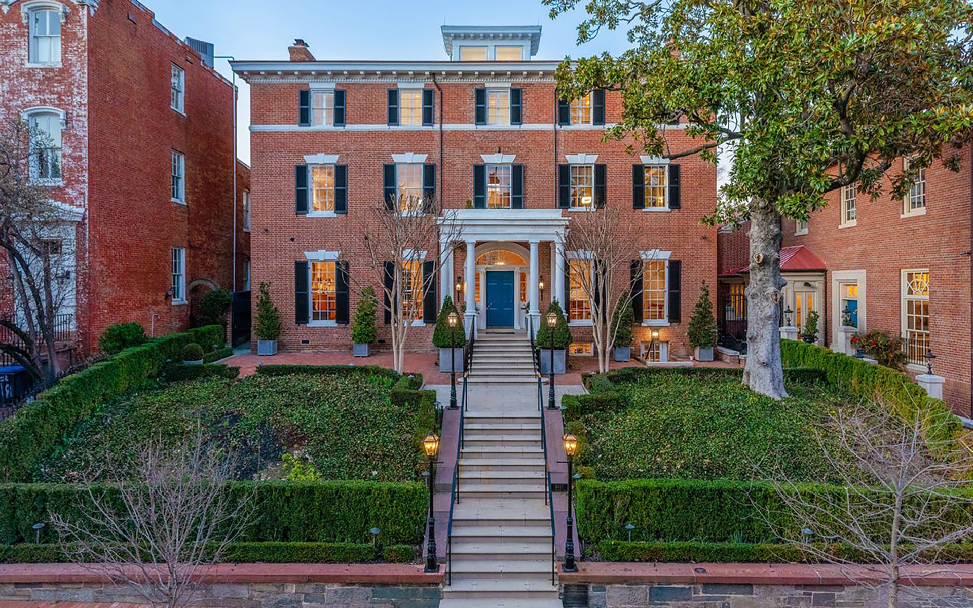 Historic brick mansion with symmetrical garden, central staircase, and large trees, set against a clear blue sky.