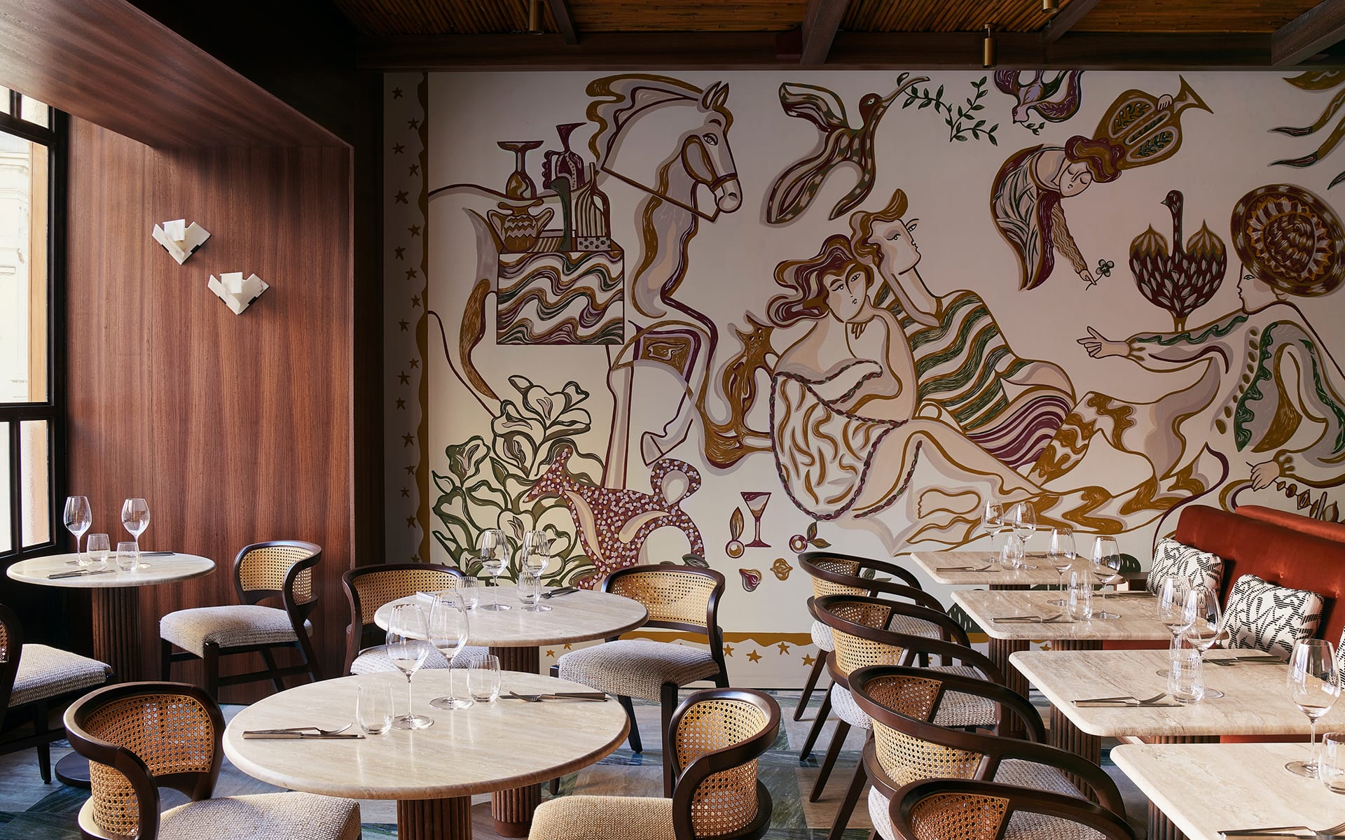 Restaurant interior with round tables, wooden chairs, and a colorful mural on the wall featuring abstract figures and patterns.