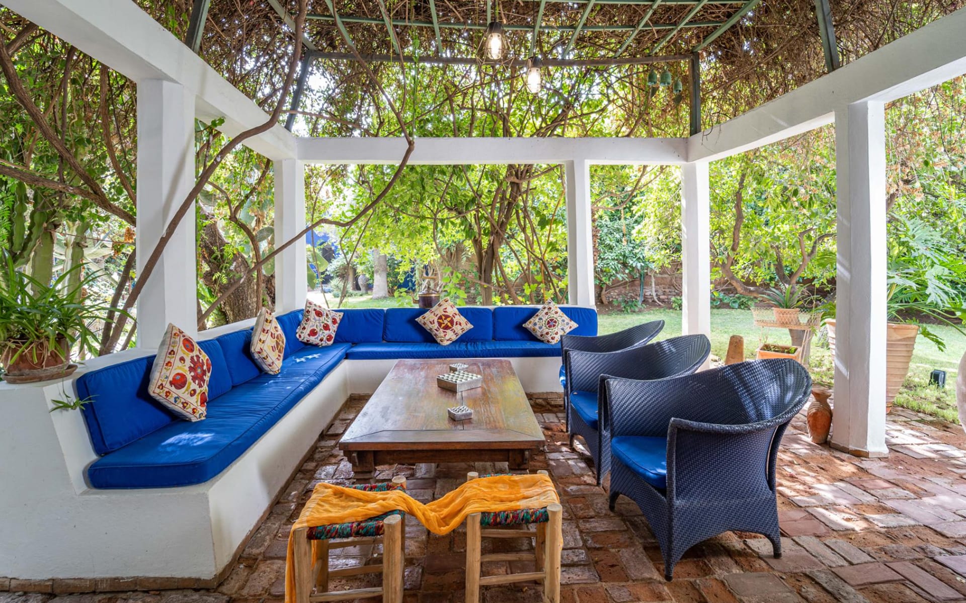 Outdoor seating area with blue cushions and colorful pillows, surrounded by greenery and a wooden table in the center.