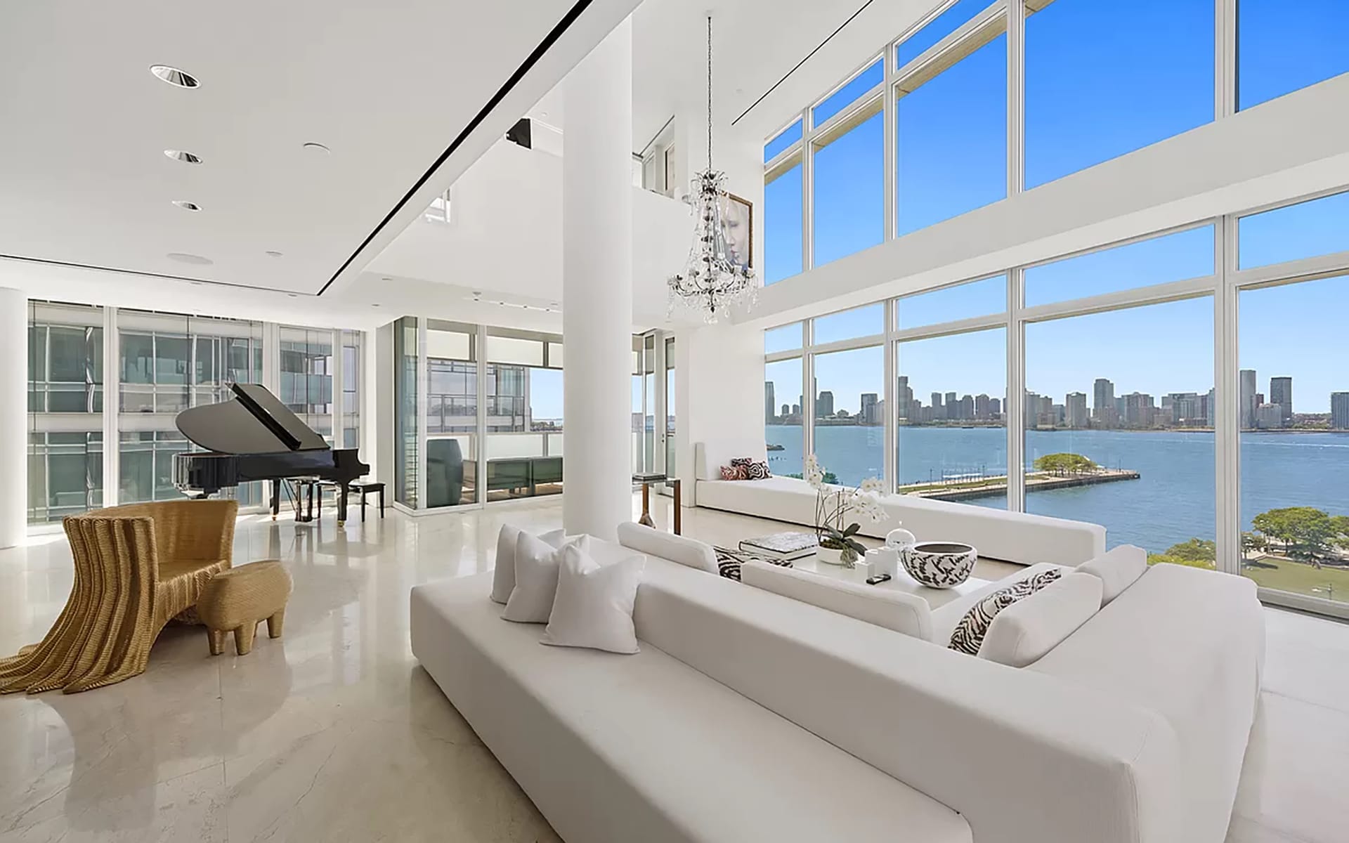 Spacious modern living room with grand piano, large windows, white furniture, city skyline, and waterfront view.