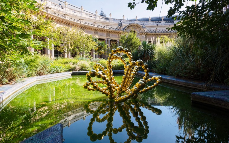 Golden bead sculpture reflecting in a pond with greenery, surrounded by a historic building in the background.