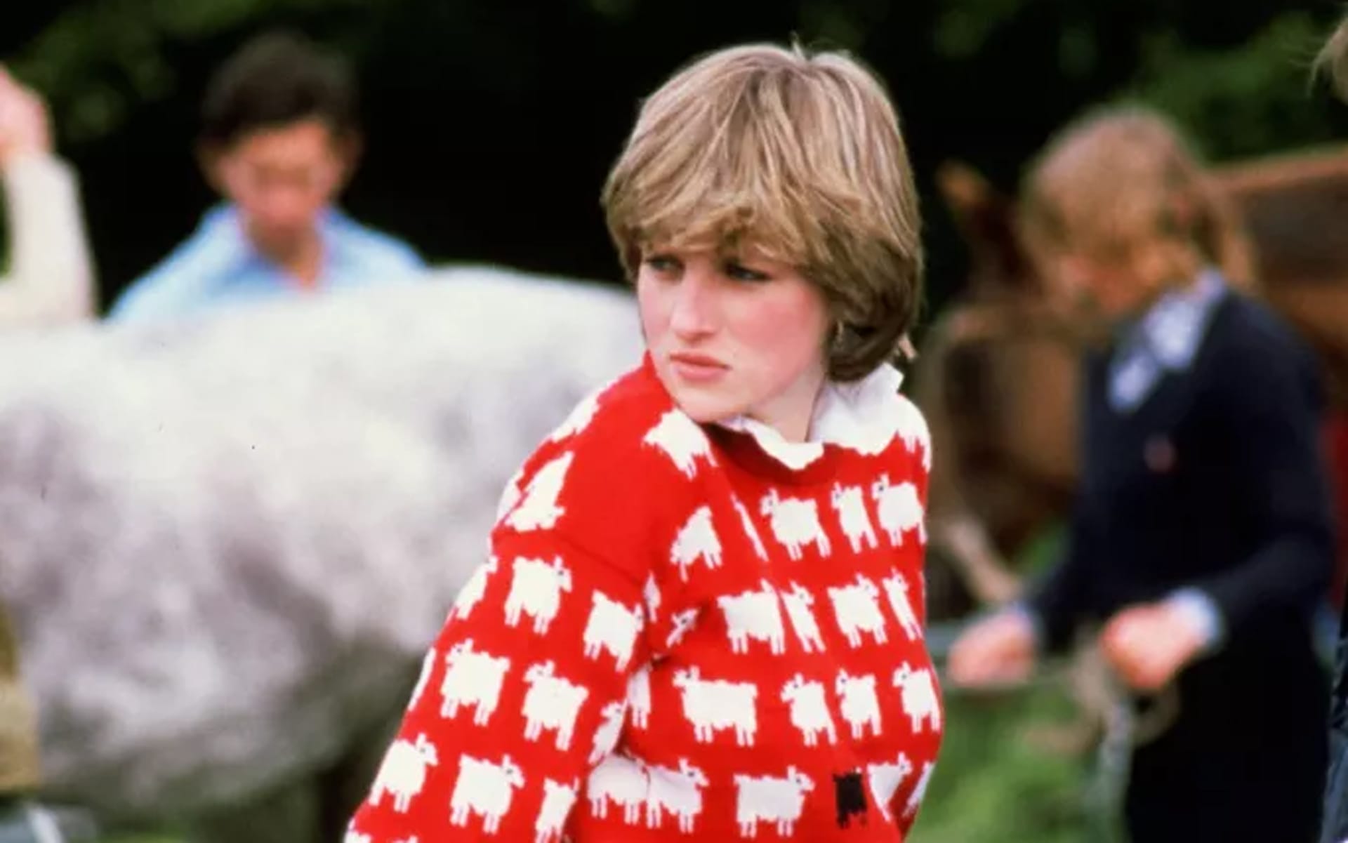 Person in a red and white sheep-patterned sweater outdoors with blurred people and a horse in the background.
