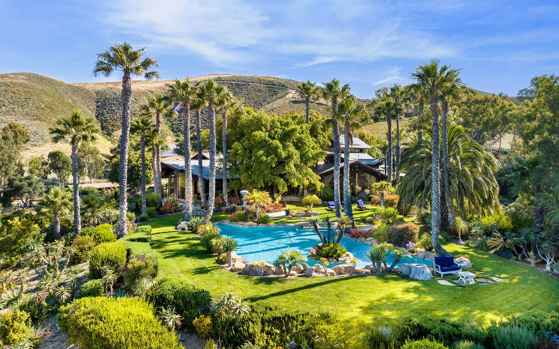 Luxurious home surrounded by lush landscaping, palm trees, and a pristine swimming pool under a clear blue sky.