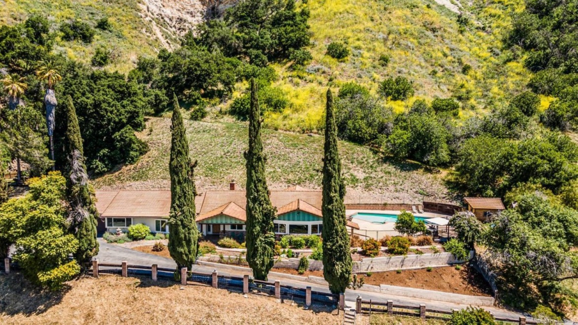 Aerial view of a hillside home with a red-tiled roof, surrounded by lush greenery and tall cypress trees.