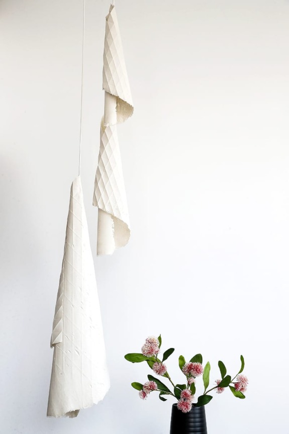 Hanging textured paper sculptures with a black vase holding pink flowers and green leaves against a plain white background.