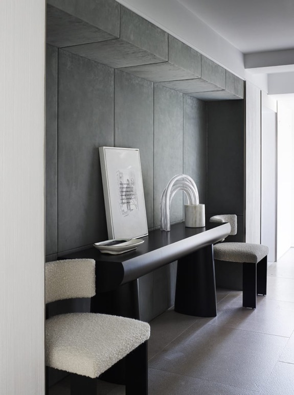 Modern hallway with dark console table, abstract art, and textured chairs under soft lighting.