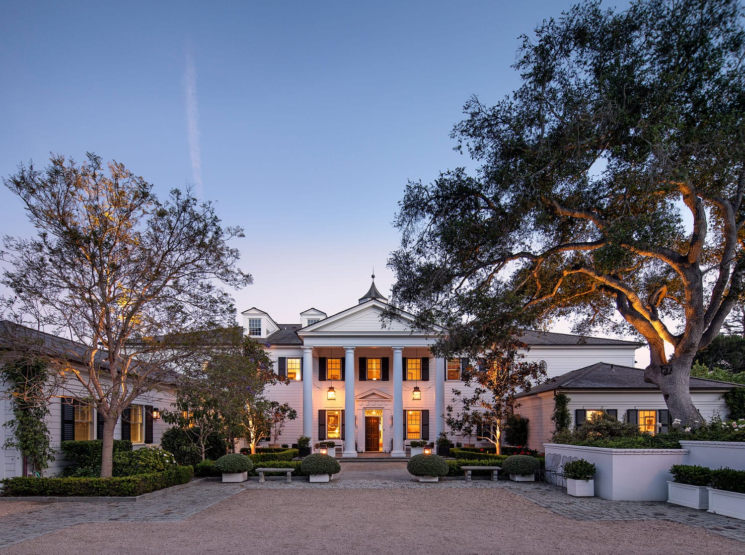 Elegant mansion with white columns and warm lighting, surrounded by large trees and a gravel driveway at dusk.