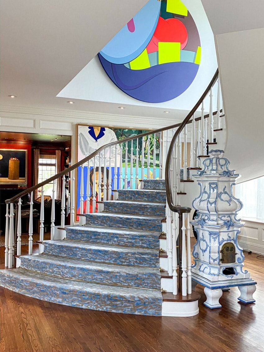 Spiral staircase with blue patterned carpet, colorful abstract art on ceiling, white walls, and decorative white fireplace.