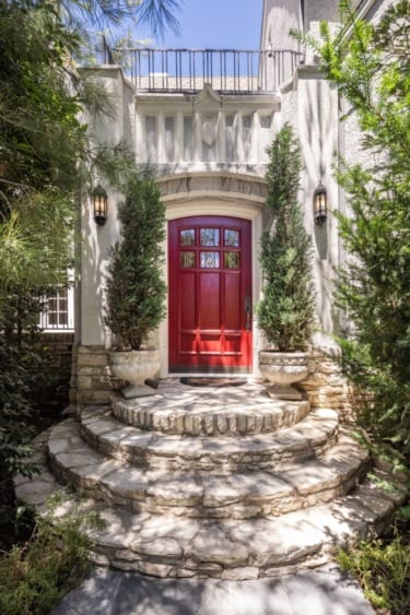 Elegant stone entrance with a vibrant red door, flanked by tall potted evergreens under a clear blue sky.