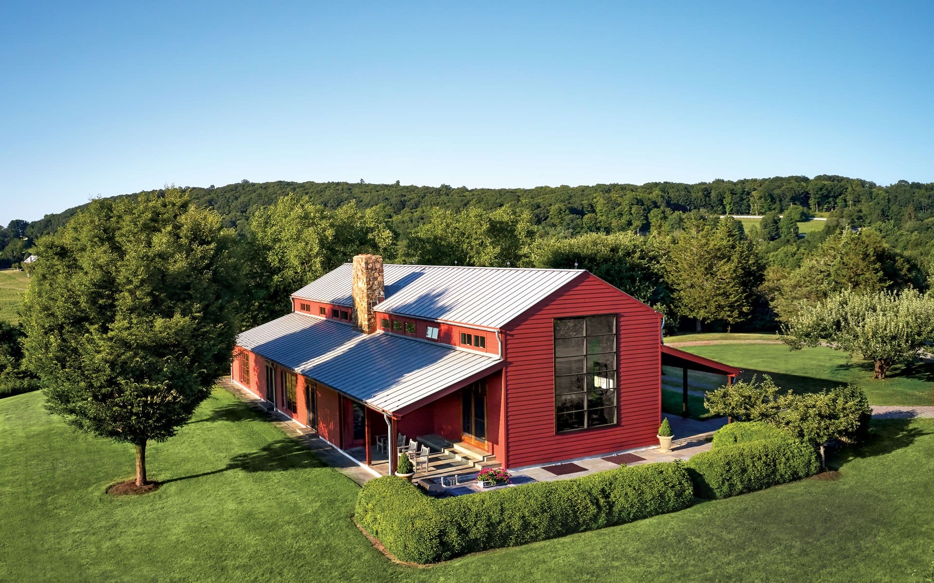 Red barn-style house with metal roof surrounded by lush green landscape and trees under a clear blue sky.