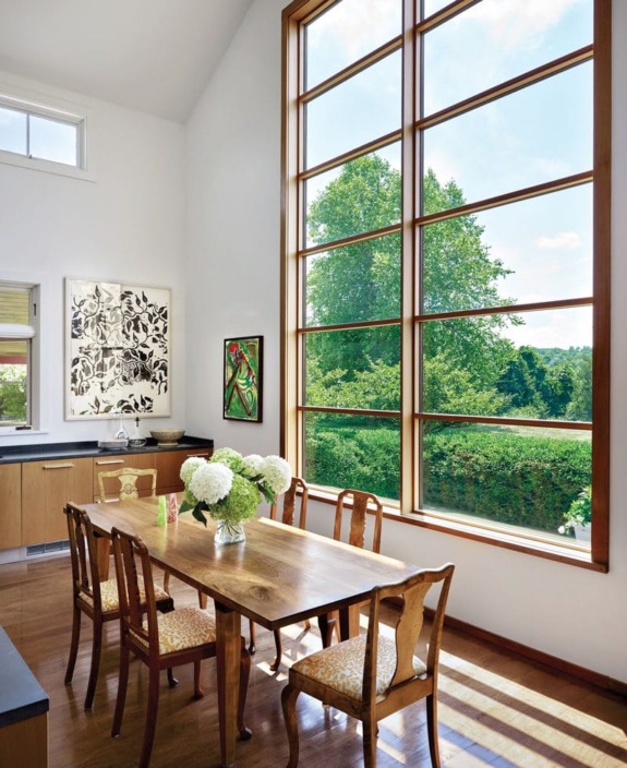 Dining room with wooden table, chairs, large window view of greenery, and modern art on white walls.