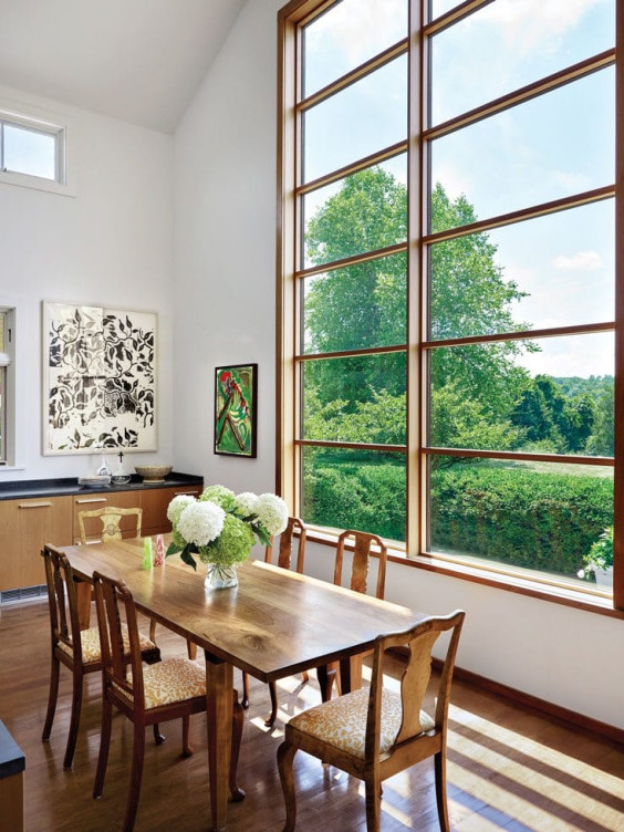 Dining room with wooden table, chairs, large window view of greenery, and modern art on white walls.